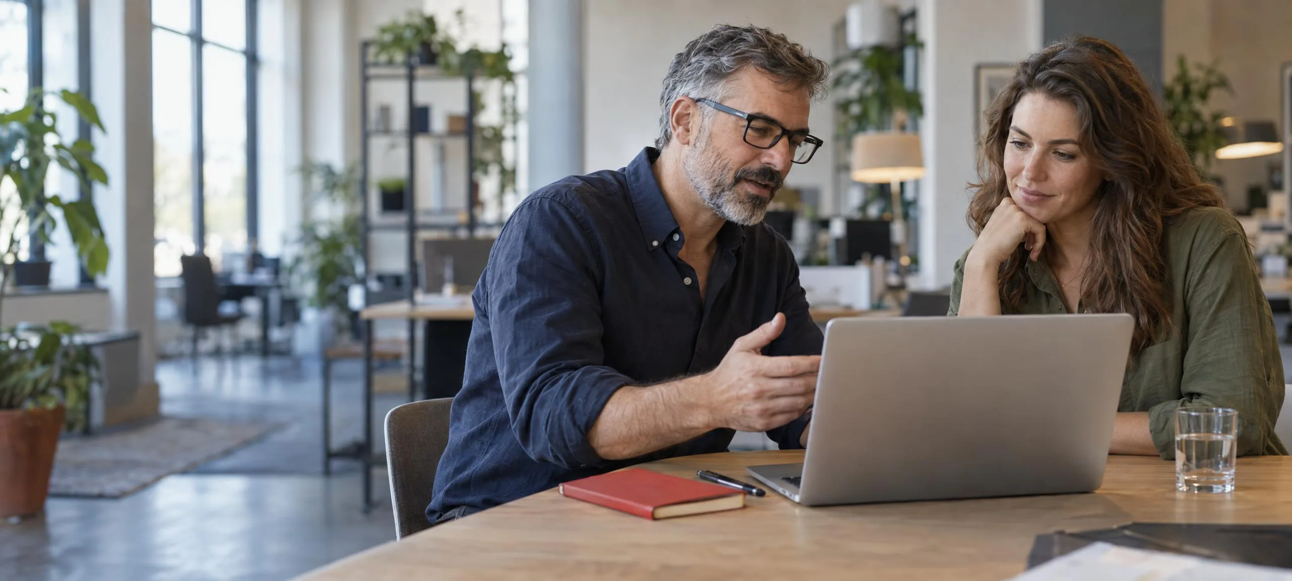 A consultant in glasses talks a business owner through a plan at a laptop in a bright open-plan office; she listens, chin on her hand.